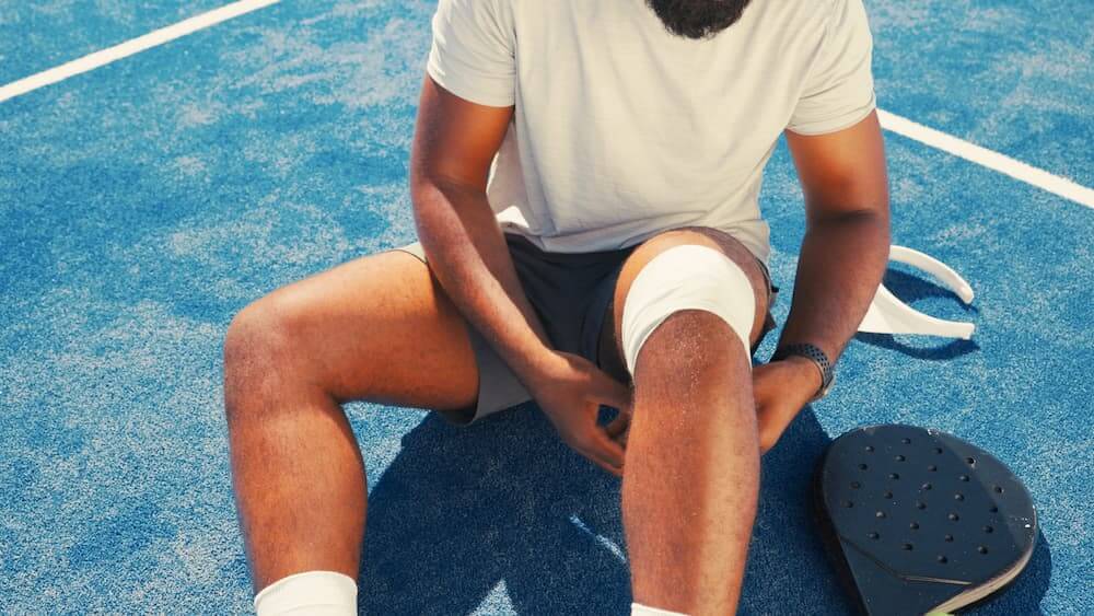 An athlete sitting on a blue court wrapping a bandage around his knee to manage overuse injuries.