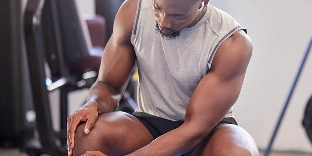 Man, fitness and knee injury in gym after workout, training or intense exercise. Earphones, health and bodybuilder male from Nigeria suffering from sore leg, muscle pain or joint inflammation.