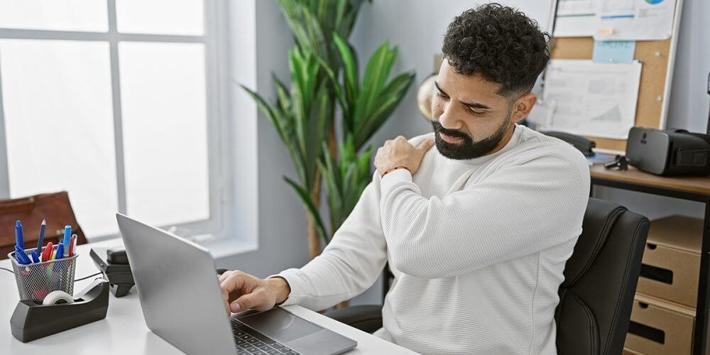 A bearded man experiences shoulder pain while working on his laptop in a bright office setting.