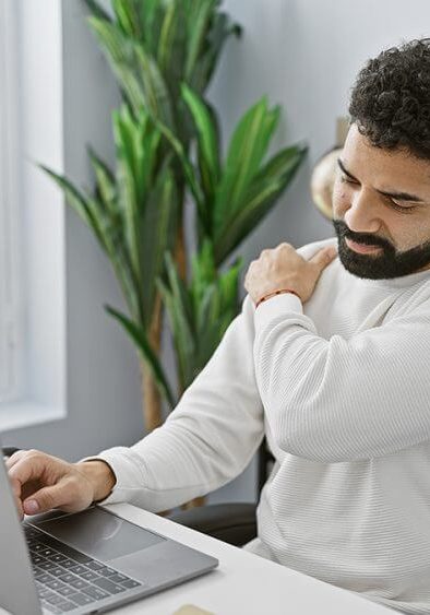 A bearded man experiences shoulder pain while working on his laptop in a bright office setting.